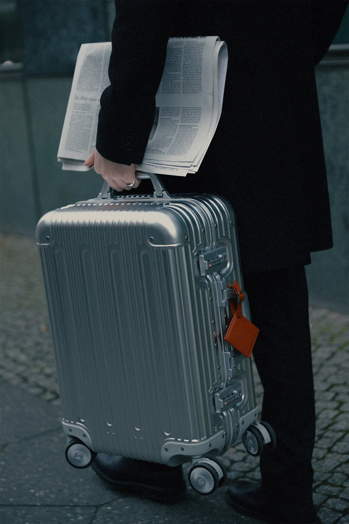 Silver PREZOR suitcase with a newspaper tucked into the handle, in the city